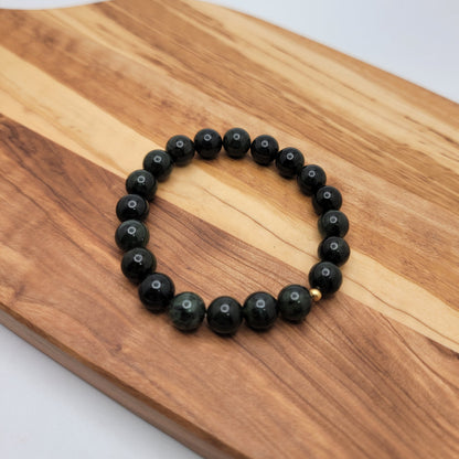 A beaded bracelet made of glossy dark green round beads, resting on a wooden board against a light gray background.