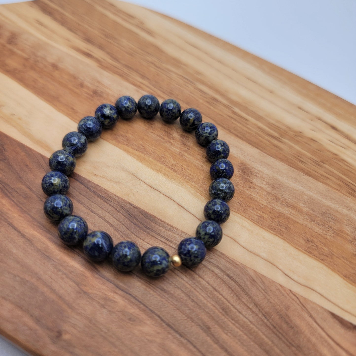 A beaded bracelet made of dark blue lapis lazuli stones with tiny gold flecks, accented by a small gold spacer bead, displayed on a wooden board.