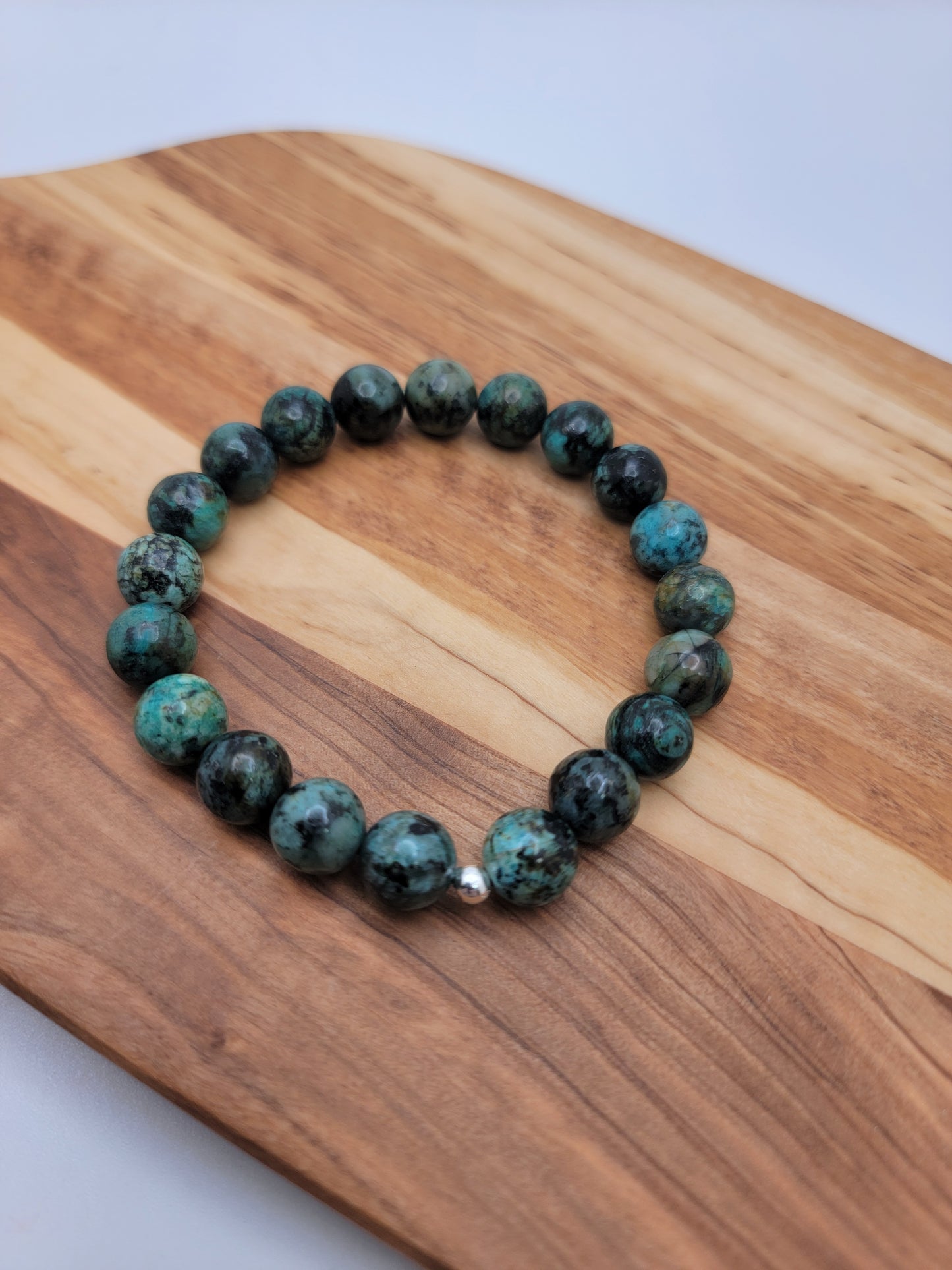 A beaded stretch bracelet made of round turquoise-green and black marbled beads with a small silver accent bead, displayed on a wooden cutting board.