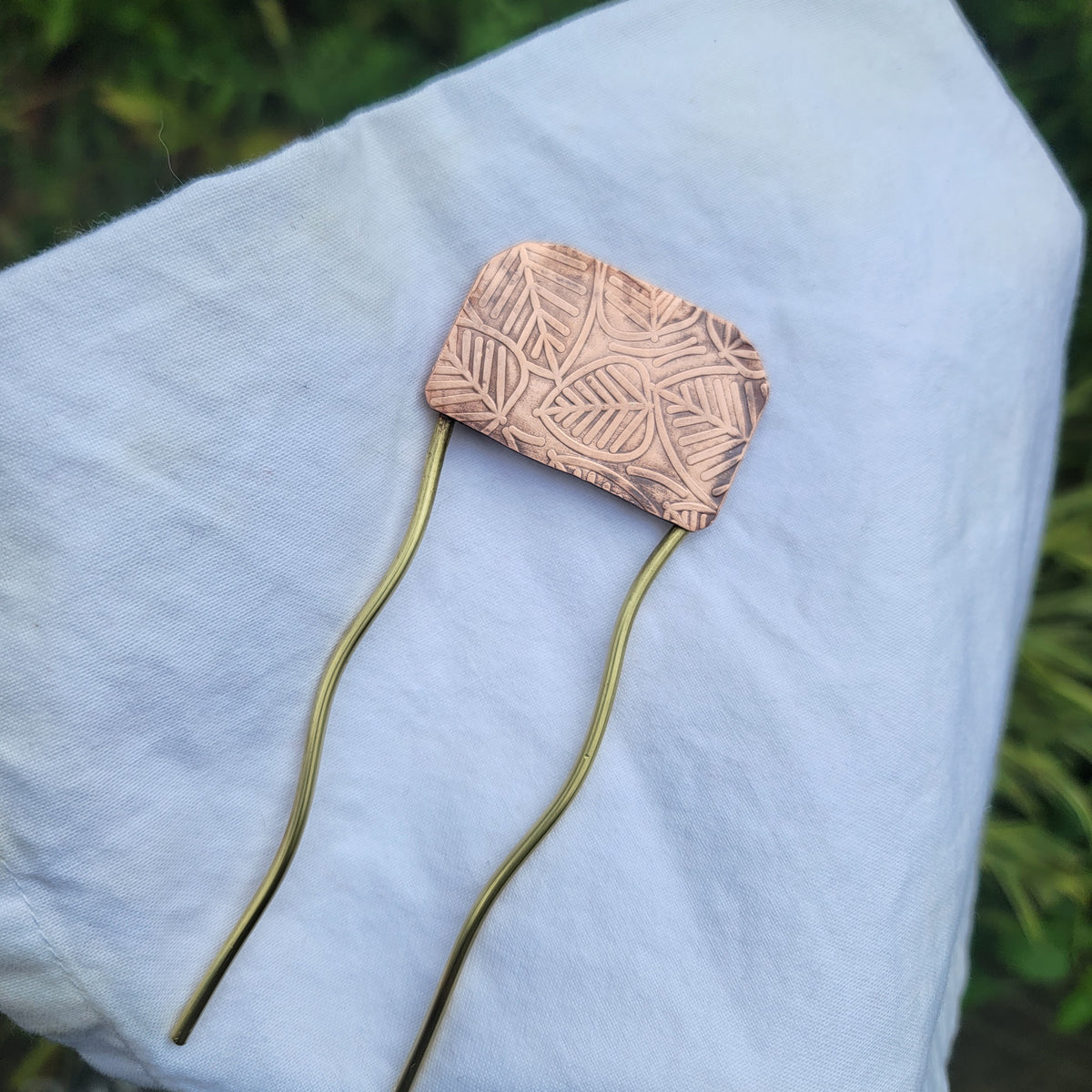 Brass and leaf patterned copper hair fork on white cloth in natural environment next to foliage