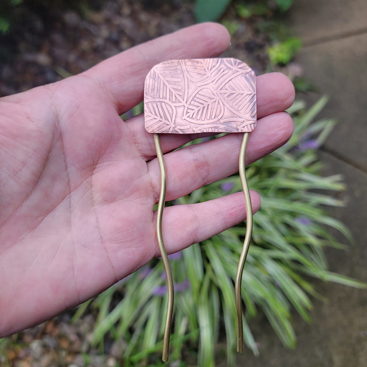 A hand holding a copper hair accessory with an engraved leaf pattern on the top and two long, wavy brass-colored metal wires extending downward, set against a blurred garden background.