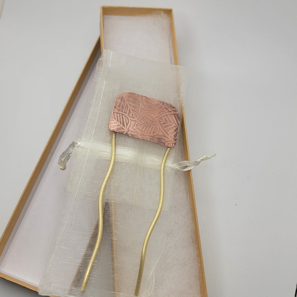 Brass and leaf patterned copper hair fork shown in organza pouch on top of gift box