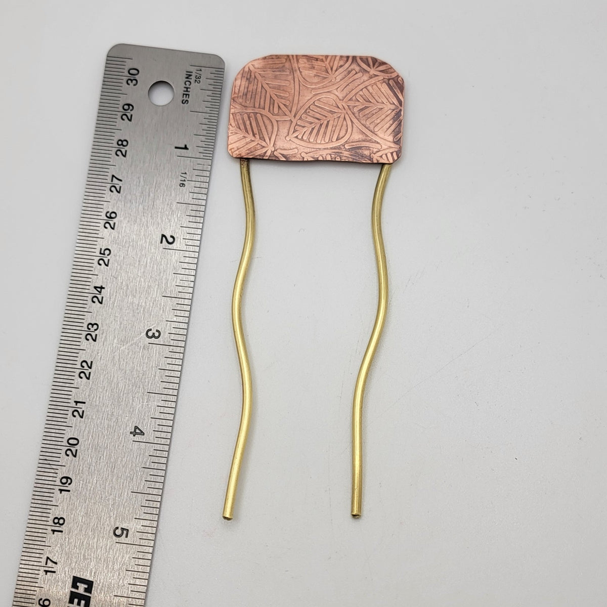 Brass and leaf patterned copper hair fork next to a ruler on a white background