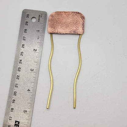 Brass and leaf patterned copper hair fork next to a ruler on a white background