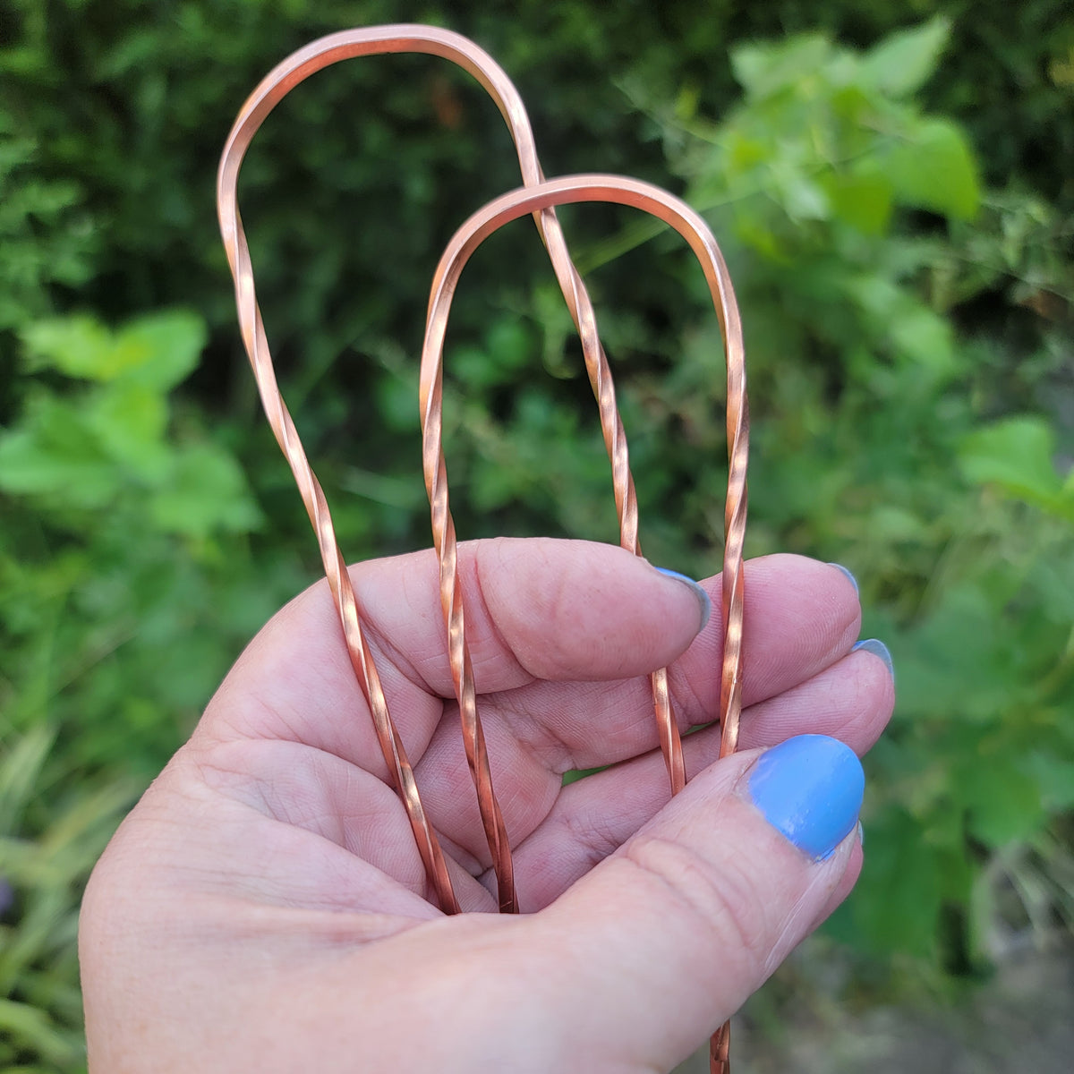 2 copper hair pins with twist design being held by a hand in natural outdoor environment