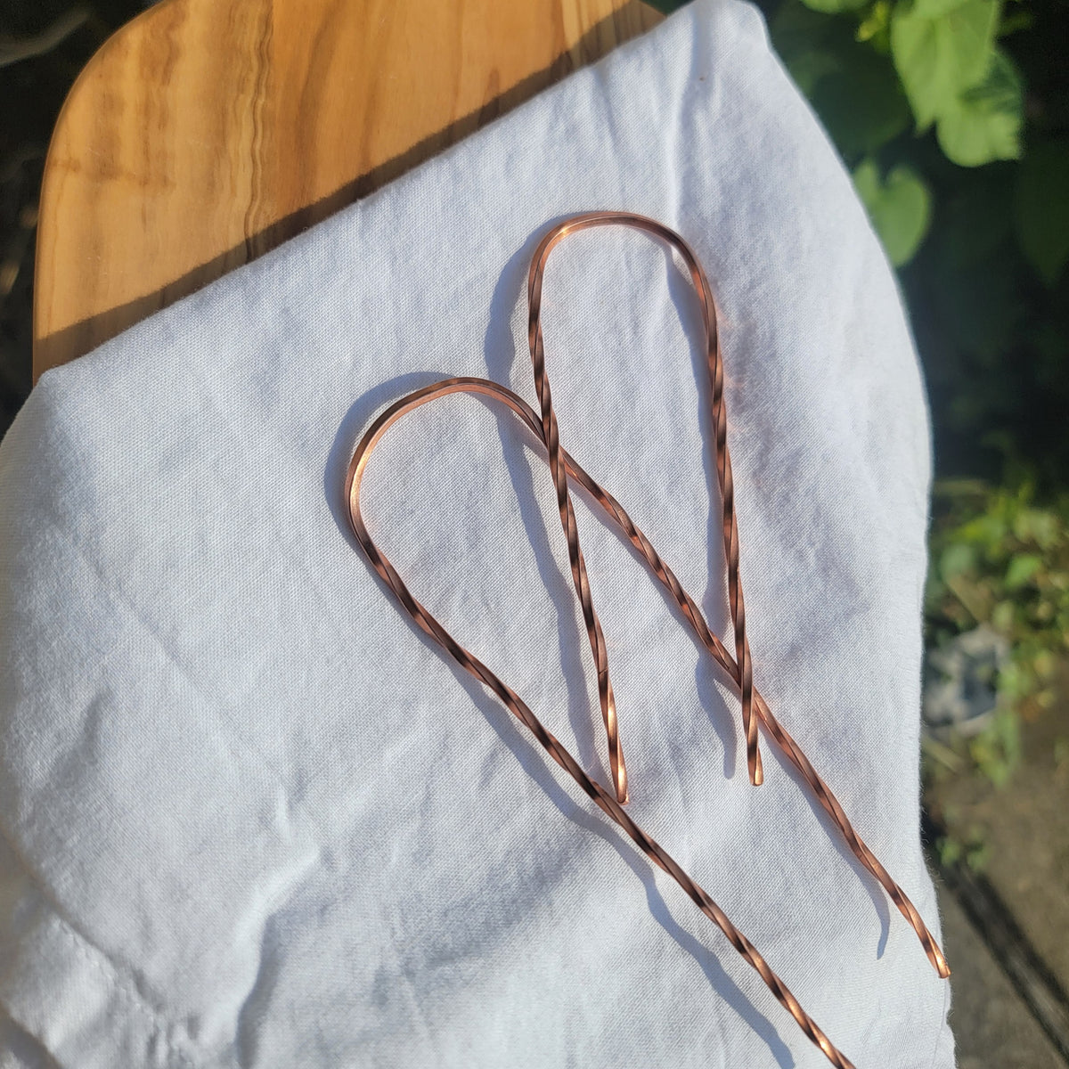 2 copper hair pins with twist design on white cloth on wood platform in natural outdoor environment