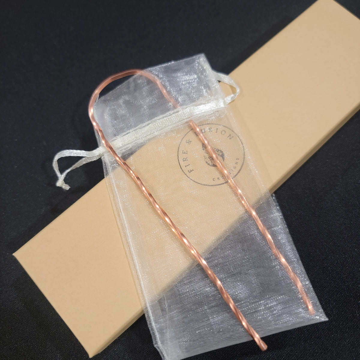 A copper-colored twisted wire hair pin in an organza drawstring pouch, placed over a tan rectangular box on a dark background.