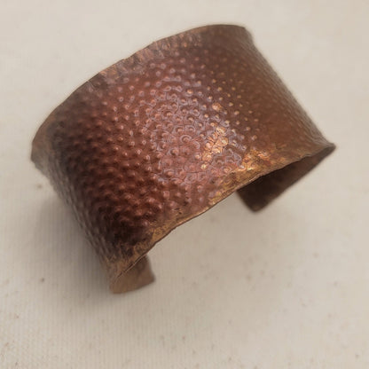 A hammered copper cuff bracelet with a textured, dimpled surface, displayed on a neutral beige fabric background.