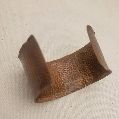 A hammered metal cuff bracelet in a warm copper-brown finish, displayed on a textured beige fabric background.