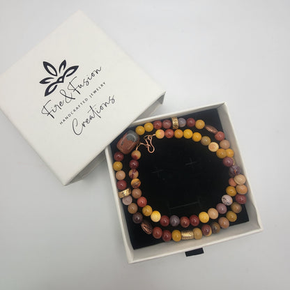 A beaded necklace displayed in an open white jewelry box next to its branded lid. The necklace features marbled beads in earthy tones of brown, yellow, orange, and purple, accented with gold-toned spacers.