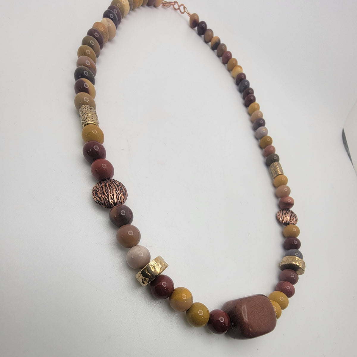 A beaded necklace featuring a mix of earthy brown, terracotta red, mustard yellow, and metallic gold beads, with a large polished reddish-brown stone pendant at the bottom center, displayed on a light gray background.
