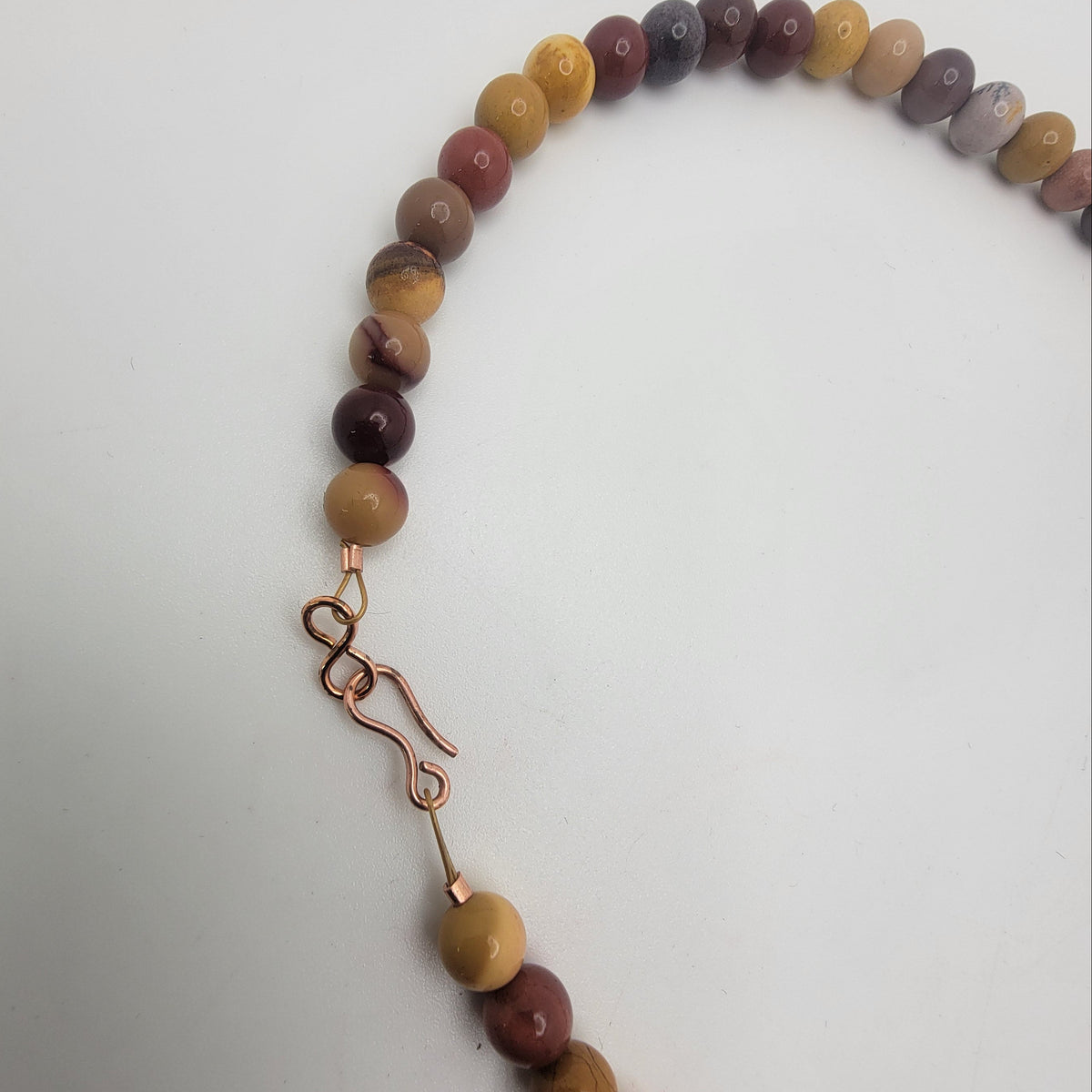 A beaded necklace made of polished round beads in earthy brown, tan, and muted purple tones, with a copper-toned metal clasp, photographed on a plain light background.