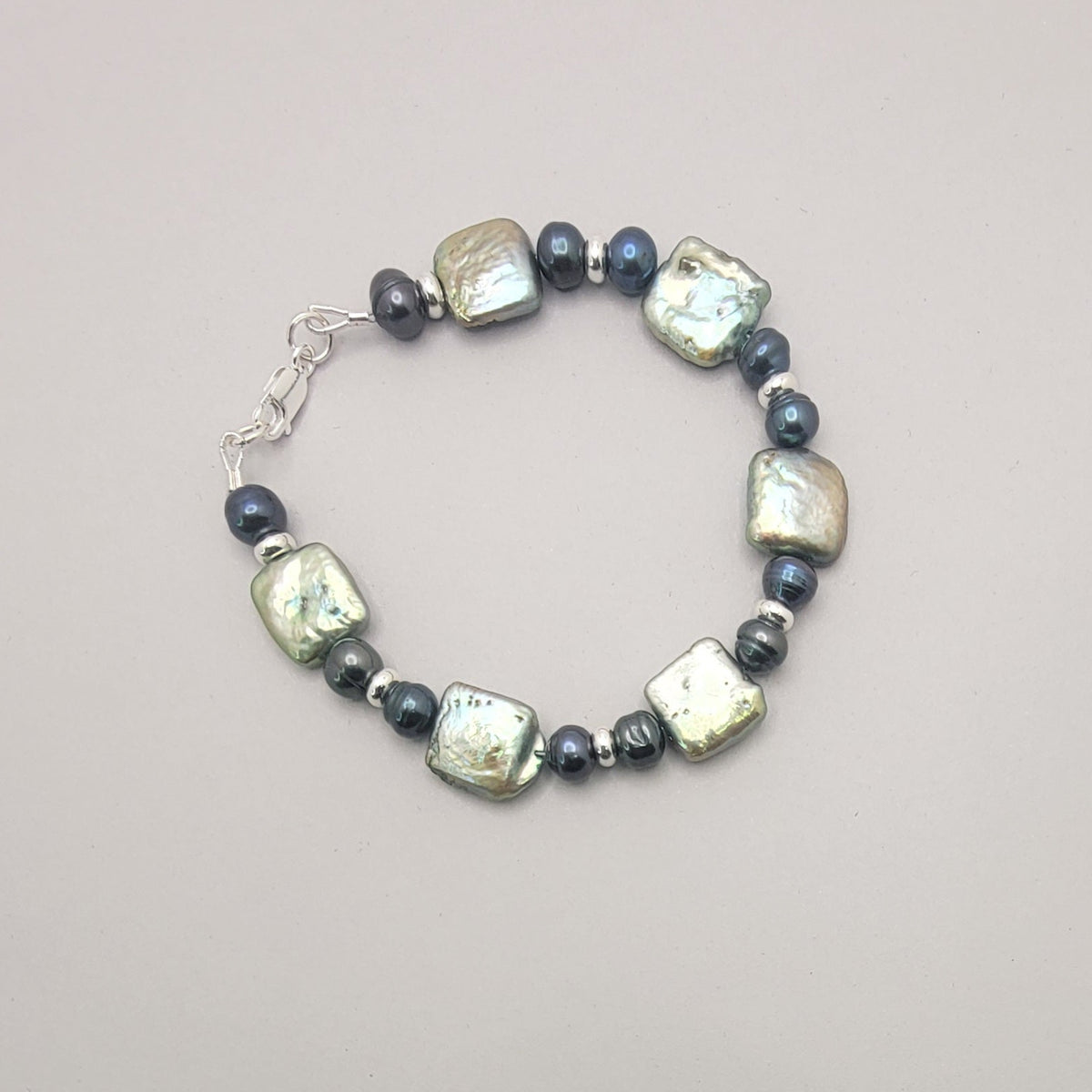 A beaded bracelet featuring baroque pearls and small dark pearls, arranged in a circle on a neutral gray background.