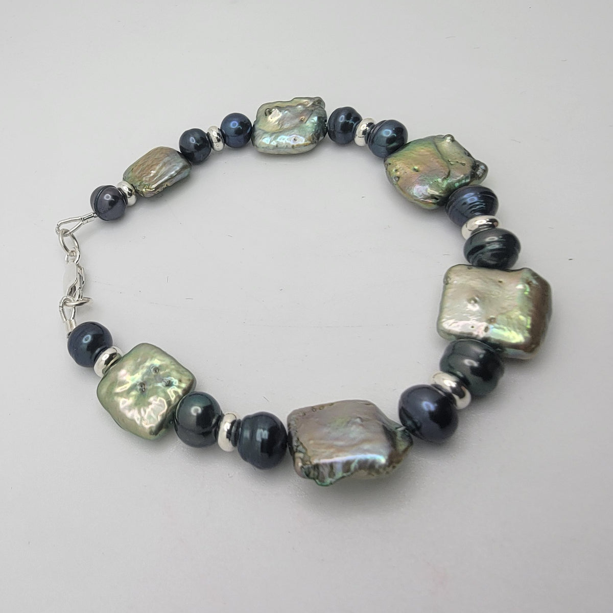 A beaded stretch bracelet featuring large, irregular baroque pearls in olive green and iridescent gray, accented by dark blue-gray beads and small silver spacer beads, with a silver clasp on a light gray background.