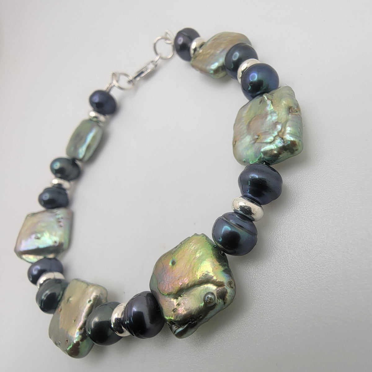 A beaded bracelet featuring large, irregular baroque pearls with iridescent green and gold hues, alternating with smooth, dark navy blue beads and small silver spacer rings, finished with a silver clasp.