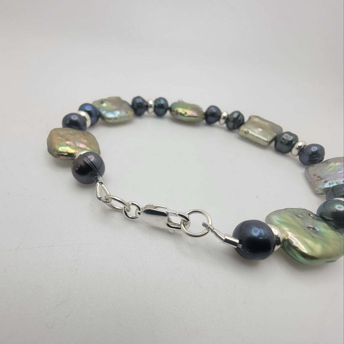 A beaded bracelet featuring large, irregular iridescent baroque pearls, small metallic silver beads, and dark gray round beads, finished with a silver lobster clasp, shown on a plain light gray background.