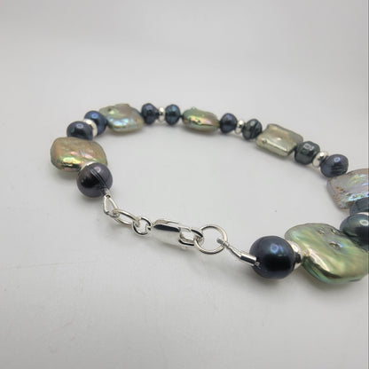 A beaded bracelet featuring large, irregular iridescent baroque pearls, small metallic silver beads, and dark gray round beads, finished with a silver lobster clasp, shown on a plain light gray background.