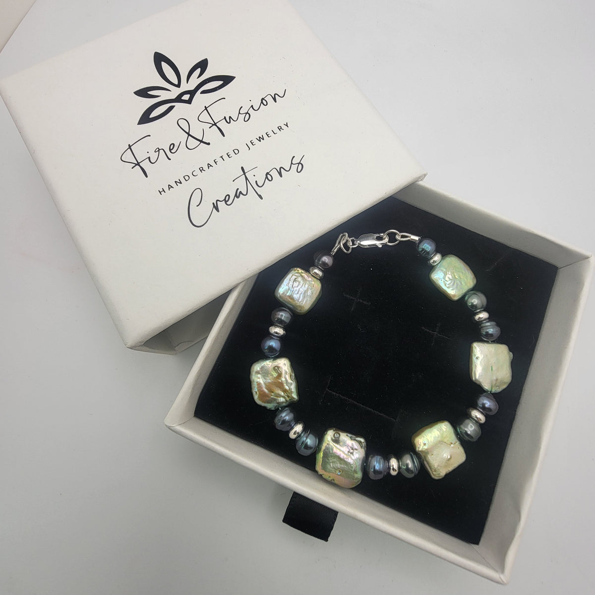 A beaded bracelet with iridescent square beads and small blue-gray beads, displayed in a black-lined gift box next to its white lid printed with the brand name 'Fire & Fusion Handcrafted Jewelry Creations'.