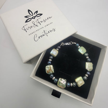A beaded bracelet with iridescent square beads and small blue-gray beads, displayed in a black-lined gift box next to its white lid printed with the brand name 'Fire & Fusion Handcrafted Jewelry Creations'.