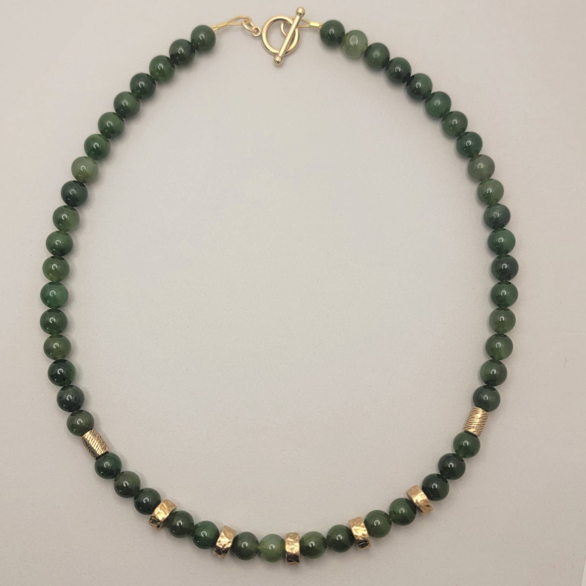 Full view of a green jade bead necklace with bronze accents and a gold-filled toggle clasp on a light background.