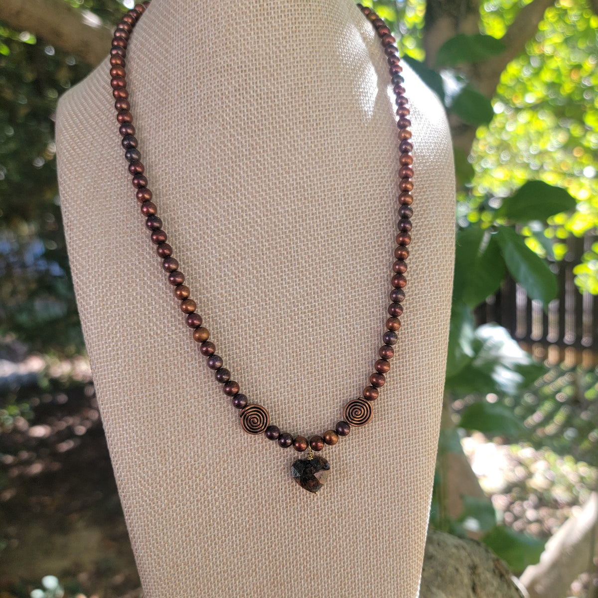 Rust pearl and feldspar heart necklace displayed on a linen bust outdoors.
