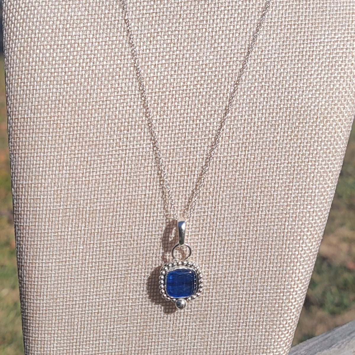 Square blue kyanite sterling silver pendant necklace displayed on a linen bust outdoors.