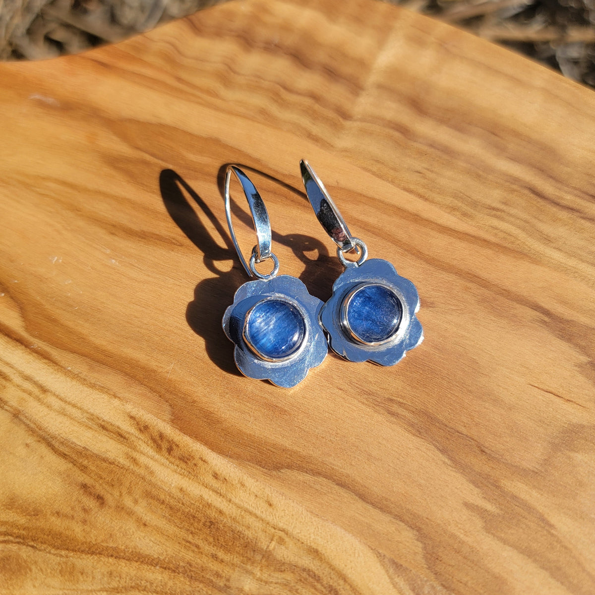 Blue kyanite earrings with scalloped sterling silver frames displayed on wood in sunlight.