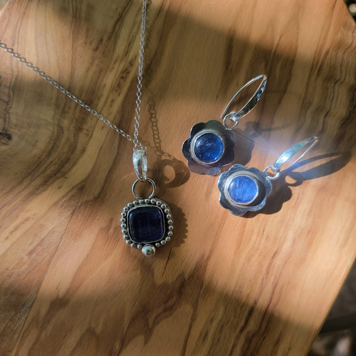 Blue kyanite earrings with scalloped sterling silver frames and a square blue kyanite  cabochon on a sterling silver chain displayed on wood in sunlight.