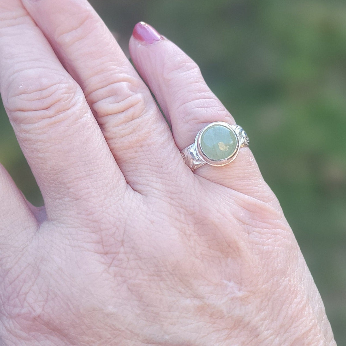 Round Aquamarine gemstone in sterling silver bezel setting with floral patterned band being shown worn on a hand in outdoor environment.