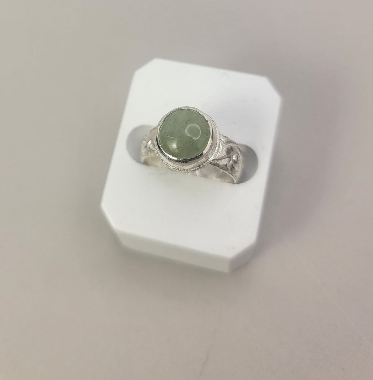 Round Aquamarine gemstone in sterling silver bezel setting with floral patterned band in white display block on neutral background.