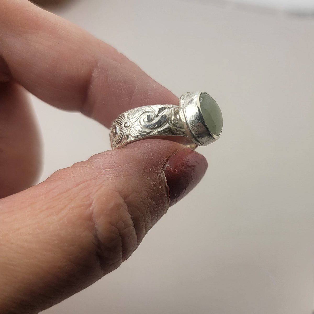 Round Aquamarine gemstone in sterling silver bezel setting with floral patterned band being held between an index finger and thumb on a neutral background.