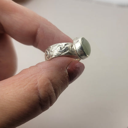 Round Aquamarine gemstone in sterling silver bezel setting with floral patterned band being held between an index finger and thumb on a neutral background.