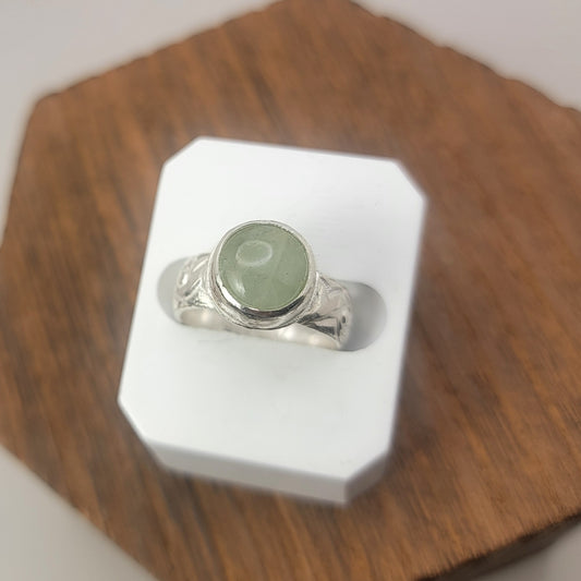 Round Aquamarine gemstone in sterling silver bezel setting with floral patterned band in white display block on a wood platform.