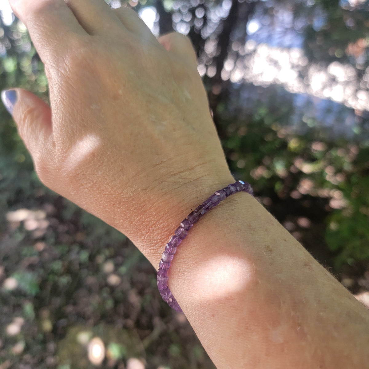 A purple beaded bracelet worn on a person's wrist, photographed outdoors with a blurred natural background of trees and sunlight.
