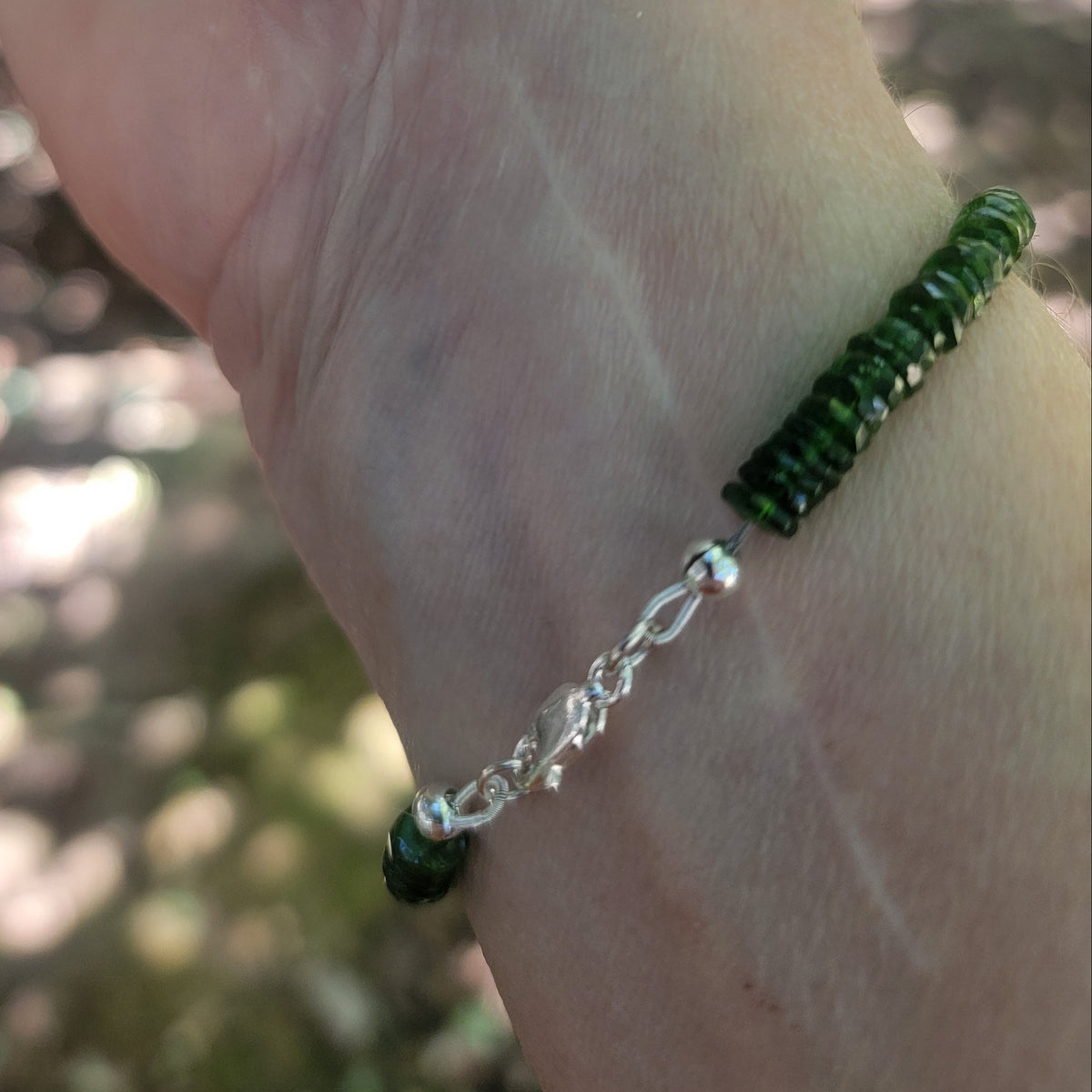 A close-up of a person's wrist wearing a beaded bracelet with dark green beads and a silver chain link accent, set against a blurred outdoor background.
