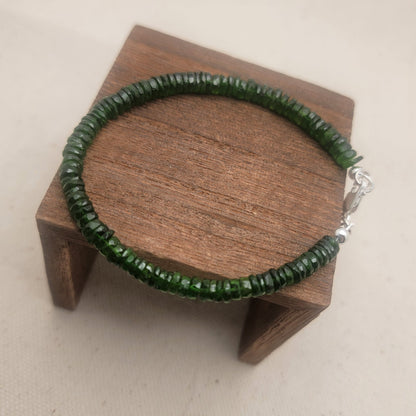 A green beaded bracelet with a silver clasp resting on a small wooden display block over a light beige fabric background.