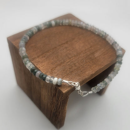 A beaded bracelet with gray, white, and dark green stones, displayed on a small wooden stand against a light gray background.