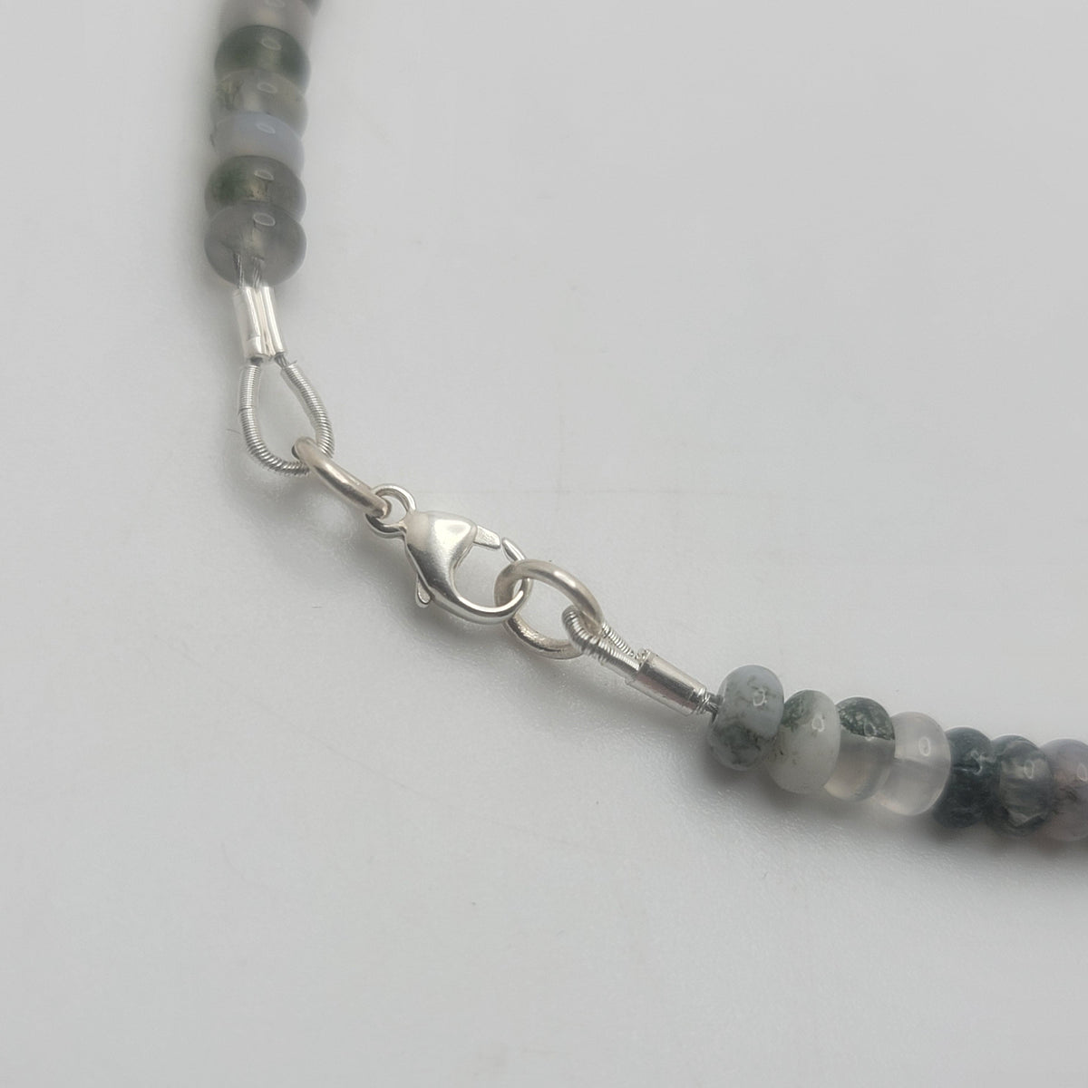 A close-up of a beaded necklace featuring round gray and white beads with a silver lobster clasp, shown on a plain light gray background.
