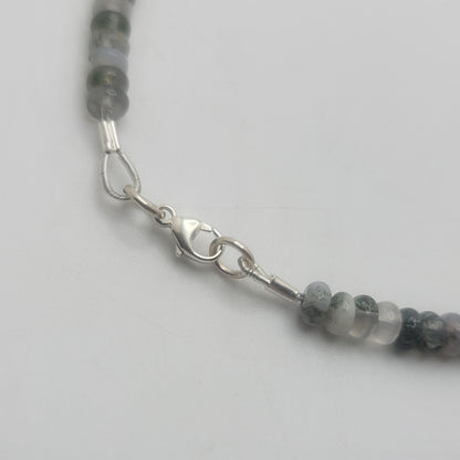 A close-up of a beaded necklace featuring round gray and white beads with a silver lobster clasp, shown on a plain light gray background.