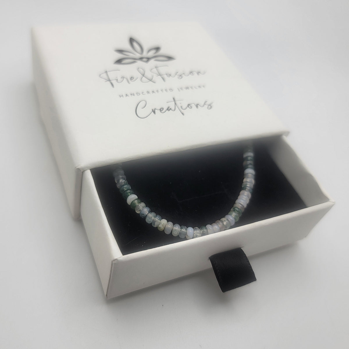 A beaded bracelet with small white and pale blue beads displayed inside an open white jewelry box. The box lid is printed with the brand name 'Fire & Fusion Creations'.