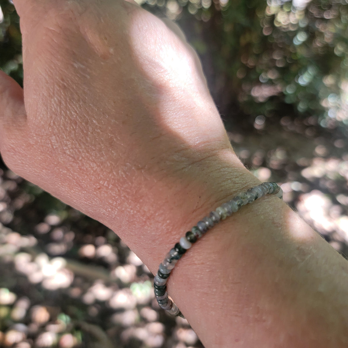 A close-up of a person's wrist wearing a beaded bracelet made of small gray and white marbled beads, photographed outdoors with a blurred natural background.