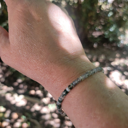A close-up of a person's wrist wearing a beaded bracelet made of small gray and white marbled beads, photographed outdoors with a blurred natural background.