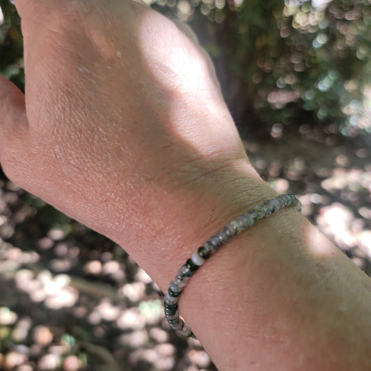 A close-up of a person's wrist wearing a beaded bracelet made of small gray and white marbled beads, photographed outdoors with a blurred natural background.