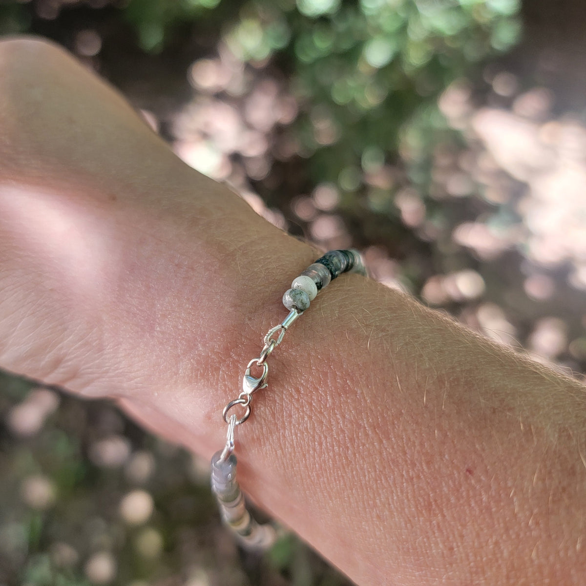 A close-up of a person's wrist wearing a beaded bracelet with silver-toned chain links and a small heart charm, set against a blurred outdoor background of green foliage.