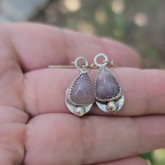 A hand holding a pair of kunzite and sterling silver earrings in a natural outdoor environment
