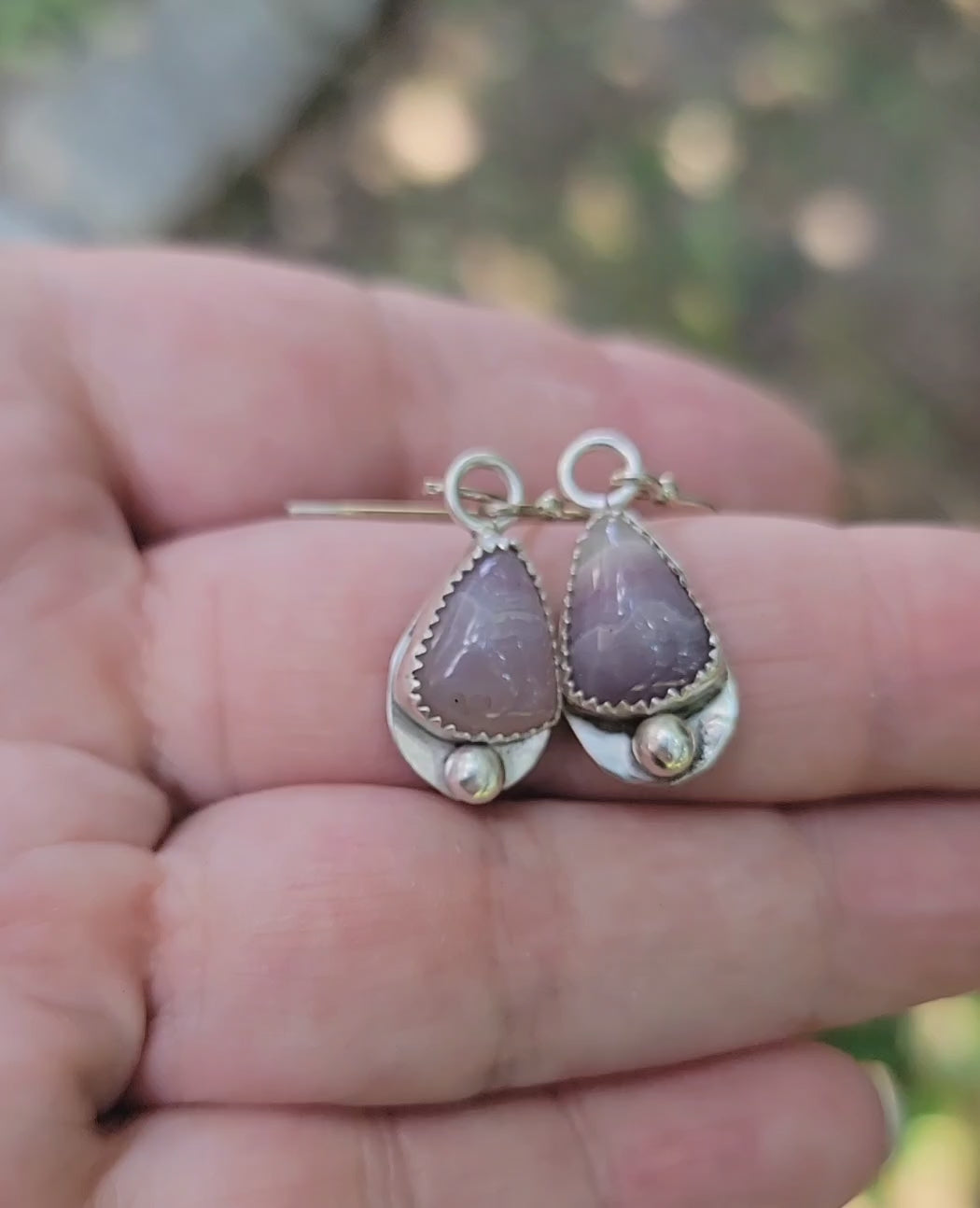 A hand holding a pair of kunzite and sterling silver earrings in a natural outdoor environment