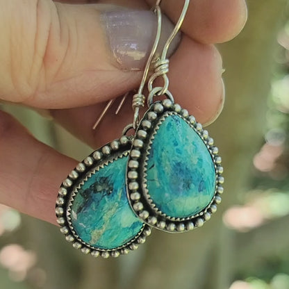 Pair of blue gemstone earrings with oxidized beaded accent on silver hooks being held in hand on a natural outdoor background