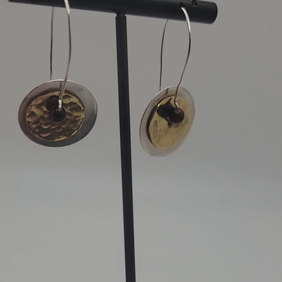 Pair of brass and silver earrings being rotated on a stand against a white background