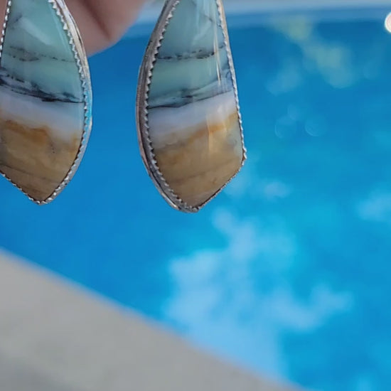 Hand holding a pair of opalized fossil cabochon earrings with sterling silver serrated bezel and smaller aquamarine accent
