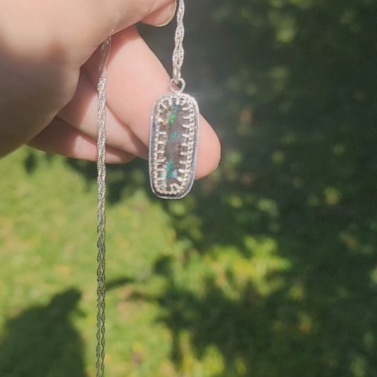 Video of hand holding iridescent pendant in natural light to show flashes of blue and green in the stone which is bezel set in sterling silver on a silver chain